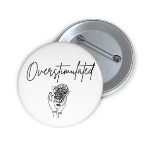 May include: White button with the word "Overstimulated" in black script. Below the text is a black line drawing of hands holding a tangled scribble. A second, silver-colored button is in the background.