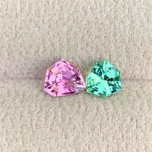 May include: Two faceted gemstones, one pink and one teal, rest on a beige surface. The gems have a triangular shape with multiple facets, catching the light. The background is blurred green foliage.