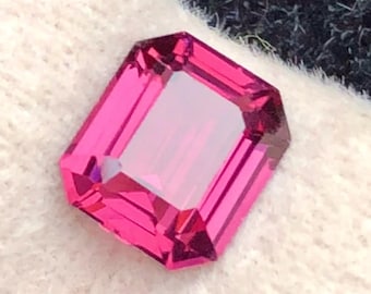 Puede incluir: Una gema rosa intenso, tallada en forma rectangular. La gema tiene un centro claro y rectangular y está colocada sobre un fondo texturizado blanco y gris oscuro. Los bordes de la gema están bien definidos.