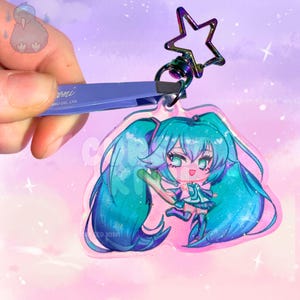 May include: A clear acrylic keychain featuring a chibi-style character with long teal hair, wearing a teal and white outfit. The charm has a glittery finish and is attached to a dark, iridescent star-shaped metal clasp. The design is transparent.