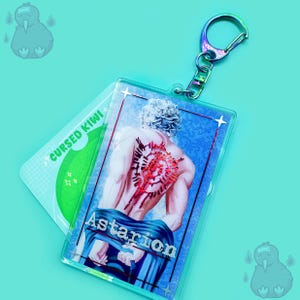Könnte beinhalten: Ein Astarion-Schlüsselanhänger mit einer Figur, die ein rotes und weißes Tattoo auf dem Rücken trägt. Der Schlüsselanhänger ist aus klarem Acryl und hat einen silbernen Metallring.