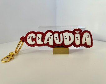 May include: Red glitter keychain with the name "CLAUDIA" in white letters and small red hearts.