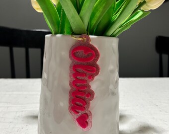 May include: A white ceramic vase with a pink glitter name tag that says "Olivia" with a pink heart at the bottom. The vase is filled with yellow tulips.