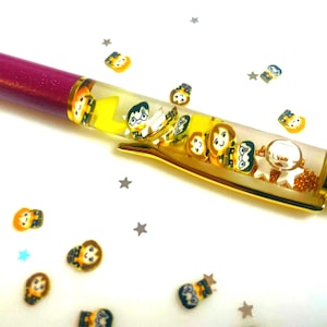 May include: A decorative pen with a burgundy barrel and gold accents. The clear barrel contains floating character figures. Small character figures and star-shaped confetti are scattered around the pen on a white surface.