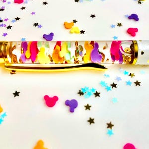 May include: A clear pen with a gold-colored clip and accents. The barrel is filled with colorful shapes and glitter. The pen's white body is decorated with small gold hearts. Scattered around the pen are star and shape-shaped confetti.