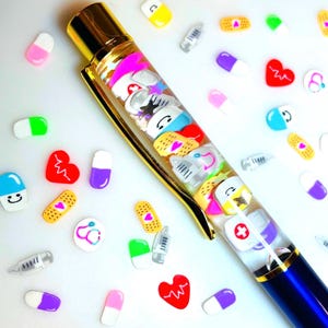 May include: A blue and gold pen filled with colorful medical-themed charms, including pills, bandages, and hearts. Scattered around the pen are additional charms of pills, bandages, and hearts. The pen has a gold clip and a blue barrel.