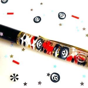 May include: A black and gold pen with a clear barrel filled with red, white, and black decorative elements. The pen is decorated with star-shaped confetti and other small shapes. The pen has a gold clip and tip.