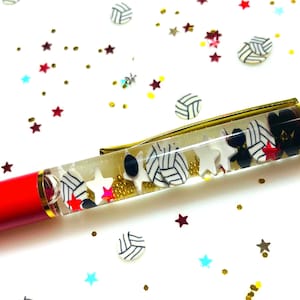 Puede incluir: Un bolígrafo rojo con clip y punta dorados, lleno de estrellas blancas, balones de voleibol blancos y negros y purpurina dorada. El bolígrafo está decorado con detalles en rojo, negro y blanco. Confeti con forma de estrella y voleibol esparcido.