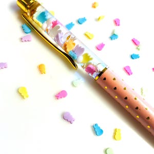 May include: A pink pen with gold accents and a clear barrel filled with colorful shapes. The pen's barrel is decorated with small gold hearts. Scattered around the pen are small, colorful shapes in various colors.