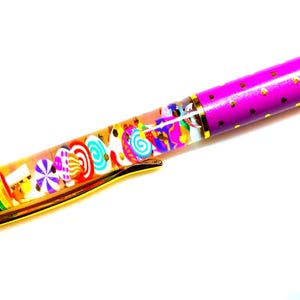 Puede incluir: Un bol&iacute;grafo colorido con un barril transparente lleno de amuletos con forma de caramelo. El bol&iacute;grafo tiene un clip dorado y un barril morado decorado con corazones dorados. El dise&ntilde;o del bol&iacute;grafo es caprichoso y divertido.