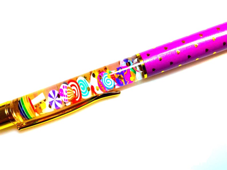 Puede incluir: Un bol&iacute;grafo colorido con un barril transparente lleno de amuletos con forma de caramelo. El bol&iacute;grafo tiene un clip dorado y un barril morado decorado con corazones dorados. El dise&ntilde;o del bol&iacute;grafo es caprichoso y divertido.