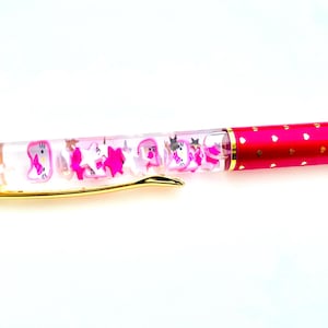 May include: A pink pen with a clear barrel filled with pink stars and unicorn figures. The pen has a gold clip and a red barrel decorated with small gold hearts. The pen is designed for writing.