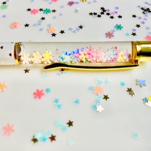 Puede incluir: Un bolígrafo blanco con detalles dorados y un clip dorado. El barril transparente está lleno de confeti colorido en forma de estrella. El bolígrafo está sobre una superficie blanca salpicada de confeti en forma de estrella de varios colores.