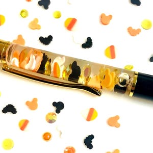 May include: A clear barrel pen with a gold clip and black end, filled with liquid and floating confetti shapes including orange, yellow, and white candy corn, black mouse silhouettes, and small yellow rhinestones, surrounded by loose confetti.