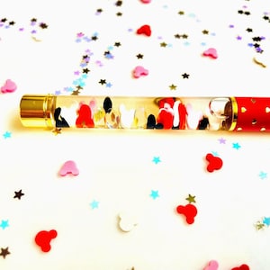 May include: A clear pen with a gold cap and red accents, filled with colorful shapes. The pen is decorated with red, white, and black shapes. Scattered around the pen are star and heart-shaped confetti in various colors.