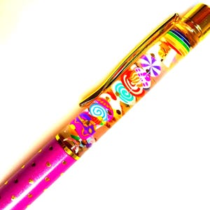 Puede incluir: Un bol&iacute;grafo decorativo con un barril transparente lleno de amuletos coloridos con forma de caramelo. El bol&iacute;grafo tiene un clip y detalles dorados, y un barril rosa con detalles dorados en forma de coraz&oacute;n. El bol&iacute;grafo est&aacute; dise&ntilde;ado para escribir.