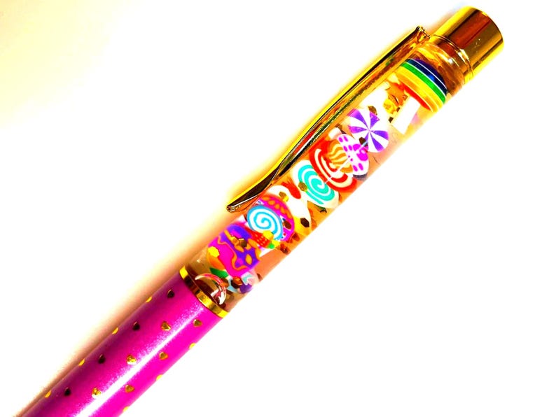 Puede incluir: Un bol&iacute;grafo decorativo con un barril transparente lleno de amuletos coloridos con forma de caramelo. El bol&iacute;grafo tiene un clip y detalles dorados, y un barril rosa con detalles dorados en forma de coraz&oacute;n. El bol&iacute;grafo est&aacute; dise&ntilde;ado para escribir.