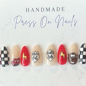 May include: A set of handmade press-on nails with a variety of designs. The nails feature red, beige, and black and white checkered patterns, Route 66 signs, and cartoon character designs. The text "HANDMADE Press On Nails" is at the top.