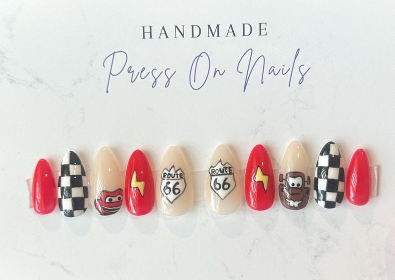 May include: A set of handmade press-on nails with a variety of designs. The nails feature red, beige, and black and white checkered patterns, Route 66 signs, and cartoon character designs. The text "HANDMADE Press On Nails" is at the top.