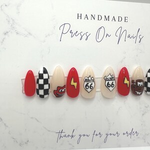 May include: Handmade press-on nails with various designs. The set includes red, black and white checkered, and beige nails. Some nails feature cartoon characters, a Route 66 sign, and a yellow lightning bolt. The card reads "Thank you for your order."