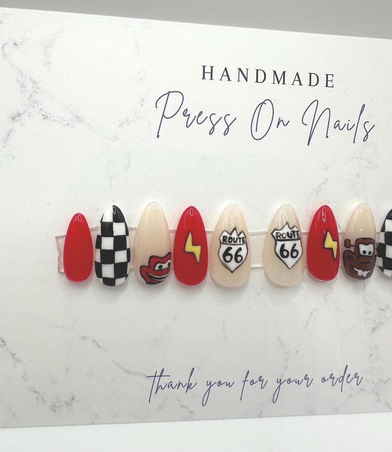 May include: Handmade press-on nails with various designs. The set includes red, black and white checkered, and beige nails. Some nails feature cartoon characters, a Route 66 sign, and a yellow lightning bolt. The card reads "Thank you for your order."