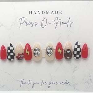 May include: A set of handmade press-on nails with a variety of designs. The nails feature red, black and white checkered patterns, cartoon characters, Route 66 signs, and lightning bolts. The card reads "Handmade Press On Nails" and "Thank you for your order."