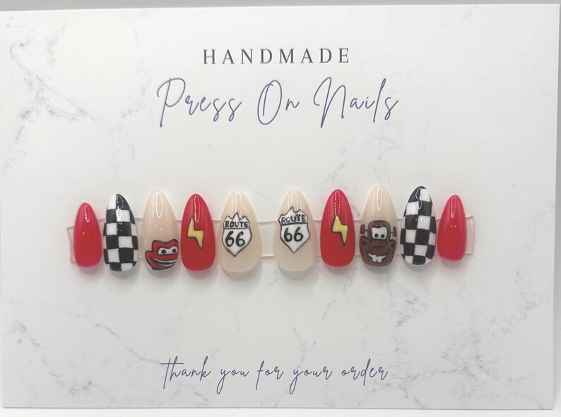 May include: A set of handmade press-on nails with a variety of designs. The nails feature red, black and white checkered patterns, cartoon characters, Route 66 signs, and lightning bolts. The card reads "Handmade Press On Nails" and "Thank you for your order."