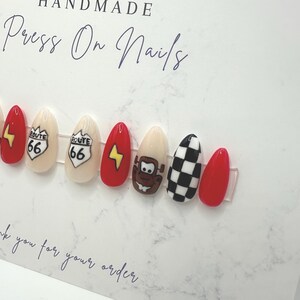 May include: A set of handmade press-on nails with various designs. The nails feature a Route 66 sign, a lightning bolt, a cartoon character, and a black and white checkered pattern. The nails are red, beige, and black.