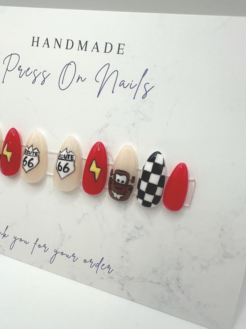 May include: A set of handmade press-on nails with various designs. The nails feature a Route 66 sign, a lightning bolt, a cartoon character, and a black and white checkered pattern. The nails are red, beige, and black.