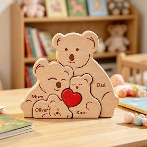Può includere: Un puzzle in legno di colore chiaro composto da quattro pezzi a forma di orso che si incastrano tra loro. I pezzi sono incisi con i nomi Mum, Dad, Oliver e Kate. Un piccolo cuore in legno rosso brillante e liscio è posizionato tra le figure.
