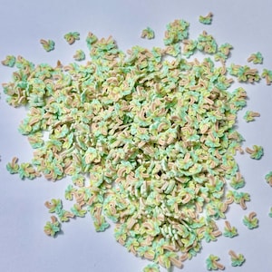 May include: A large collection of small, flat, palm tree-shaped confetti pieces. Each piece features a light mint green palm frond top and a pale peach-colored trunk. The palm tree shapes are scattered across the surface in a dense, irregular pile.