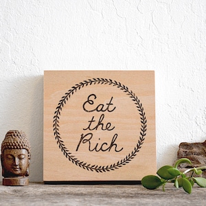 Puede incluir: Un cuadrado de madera con la frase "Eat the Rich" en un diseño de corona circular. A la izquierda del letrero hay una pequeña estatua de Buda y una piña. A la derecha hay un trozo de madera flotante y una pequeña planta.