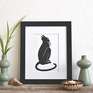 May include: Black and white silhouette of a cat sitting in a black frame with a white mat. The cat is facing to the right and looking up. The frame is on a wooden surface with a green vase and a woven basket.