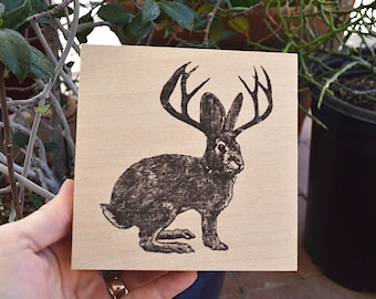 May include: A black and white illustration of a rabbit with large antlers on a wooden background.