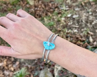 May include: A silver cuff bracelet with a turquoise stone in the center. The turquoise stone is heart-shaped and set in a silver bezel. The bracelet has a textured design.