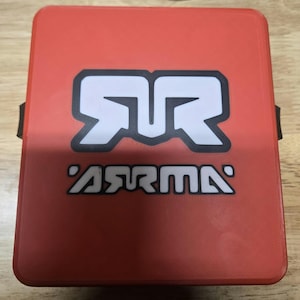 Peut inclure: Un objet carré rouge avec les lettres blanches "RR" et le mot "ARMA" en texte blanc. Les lettres sont soulignées en noir. L'objet a une sangle noire sur le côté.
