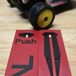 Può includere: Un cartello rosso stampato in 3D con la parola "Push" e una freccia, accanto a un componente nero. Un'auto telecomandata argento e nera è sullo sfondo.
