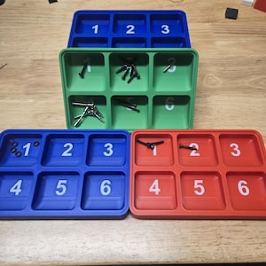 Puede incluir: Tres bandejas multicompartimento en azul, verde y rojo. Cada bandeja tiene seis compartimentos numerados. Las bandejas contienen varios artículos de ferretería pequeños. Los números 1, 2, 3, 4, 5 y 6 están impresos en blanco.