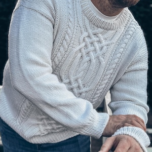Puede incluir: Un suéter de punto de cable color crema con cuello redondo y mangas largas. El suéter presenta intrincados patrones de cable en el pecho y las mangas. La persona lleva vaqueros azules. Una prenda clásica y cálida.