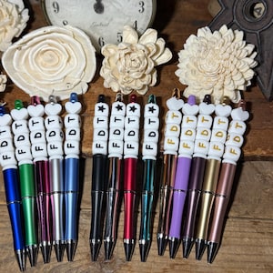 May include: Twelve pens with white beads spelling out "FDT" in gold letters. The pens are in various colors, including blue, green, red, silver, and gold.