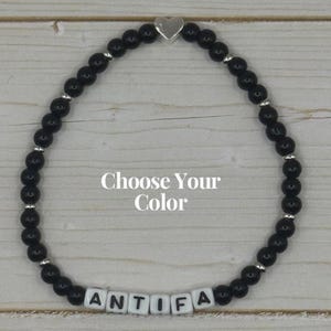 May include: Black beaded bracelet with silver accents and a silver heart charm. The bracelet spells out "ANTIFA" with white letter beads. The text "Choose Your Color" is also visible.