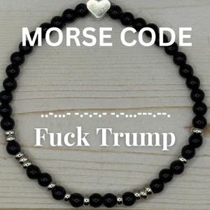May include: A circular beaded bracelet featuring small, glossy black beads interspersed with metallic silver-toned spacer beads and a single silver heart-shaped charm at the top. The text MORSE CODE and FUCK TRUMP appear with Morse code dots and dashes.