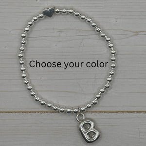 May include: Silver beaded bracelet with a heart charm and a letter "B" charm. The bracelet is strung with small, round beads. The text "Choose your color" is displayed on the image.
