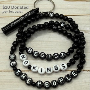 May include: Black beaded bracelets with white letter beads spelling "RESIST", "NO KINGS", and "THE PEOPLE". A black keychain with the words "Abolish ICE" is attached. The bracelets are adorned with star-shaped beads.