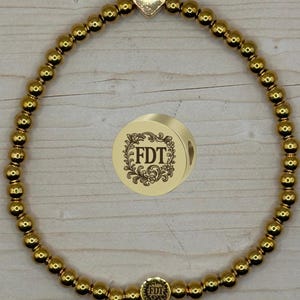 May include: Gold beaded bracelet with a heart-shaped charm and a round charm with the initials "FDT" in a floral design. The bracelet is made of small, round gold beads.