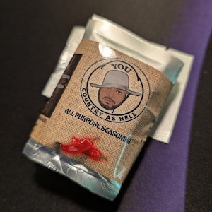 May include: A small, silver foil packet of "You Country As Hell" all-purpose seasoning. The packet features a cartoon illustration of a man wearing a hat and sunglasses. The text "You Country As Hell" is in a circle around the man's head. The packet also features three red chili peppers.