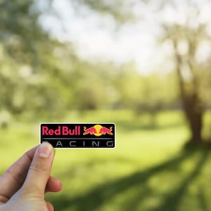 以下が含まれることがあります： 「Red Bull Racing」というテキストと赤い雄牛のロゴが入った黒と赤のステッカーです。
