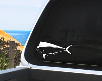 May include: A white vinyl decal of a fish, likely a mahi-mahi, is affixed to a vehicle window. The decal is a simplified, stylized illustration of the fish, with a prominent dorsal fin and a curved tail. The background shows a coastal scene.