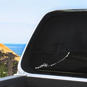 May include: A white vinyl decal of the Florida Keys on the back window of a pickup truck. The truck is parked in front of a beach scene with blue water and a sandy shore.