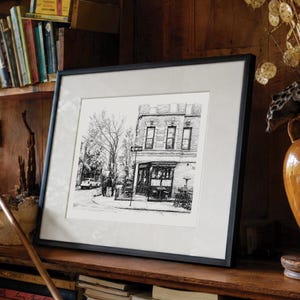 May include: A black and white ink drawing of a city street corner featuring a multi-story building with large windows, a street sign, and outdoor seating, presented in a black frame with a wide white mat board border for a clean display.